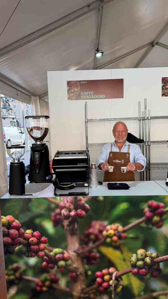 Un barista sorridente al bancone di un caffè, con attrezzature da caffè professionali e una pianta di caffè con bacche colorate in primo piano.