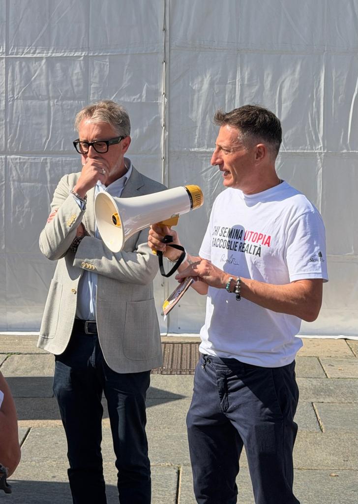 Due uomini in un evento all'aperto, uno con un megafono mentre parla e l'altro in ascolto, con una dozzina di persone che li osservano.