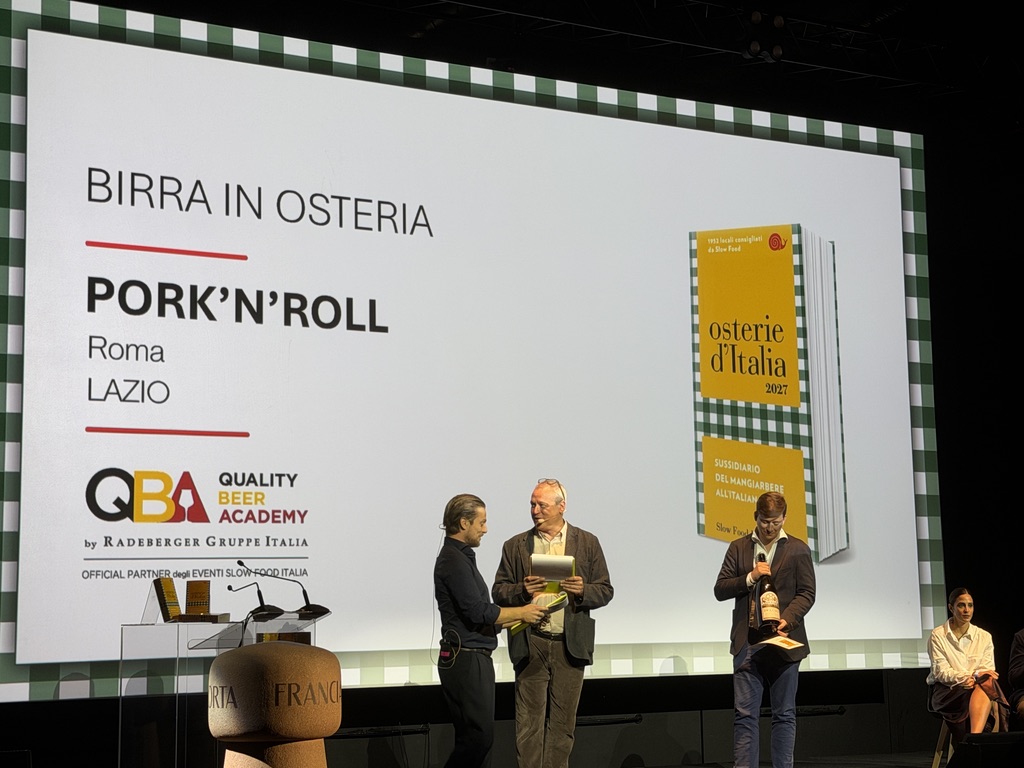 Presentazione del libro 'Osterie d'Italia 2027' con relatori che discutono davanti a uno schermo. Il titolo evidenzia 'Birra in Osteria' e 'Pork'n'Roll' a Roma. Sul palco ci sono più persone, mentre nella parte sinistra si vede un tavolo con attrezzature.